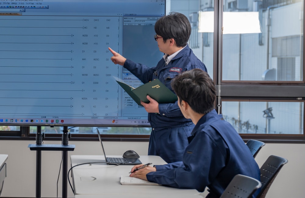 モニターを指しながら同僚と打ち合わせる社員